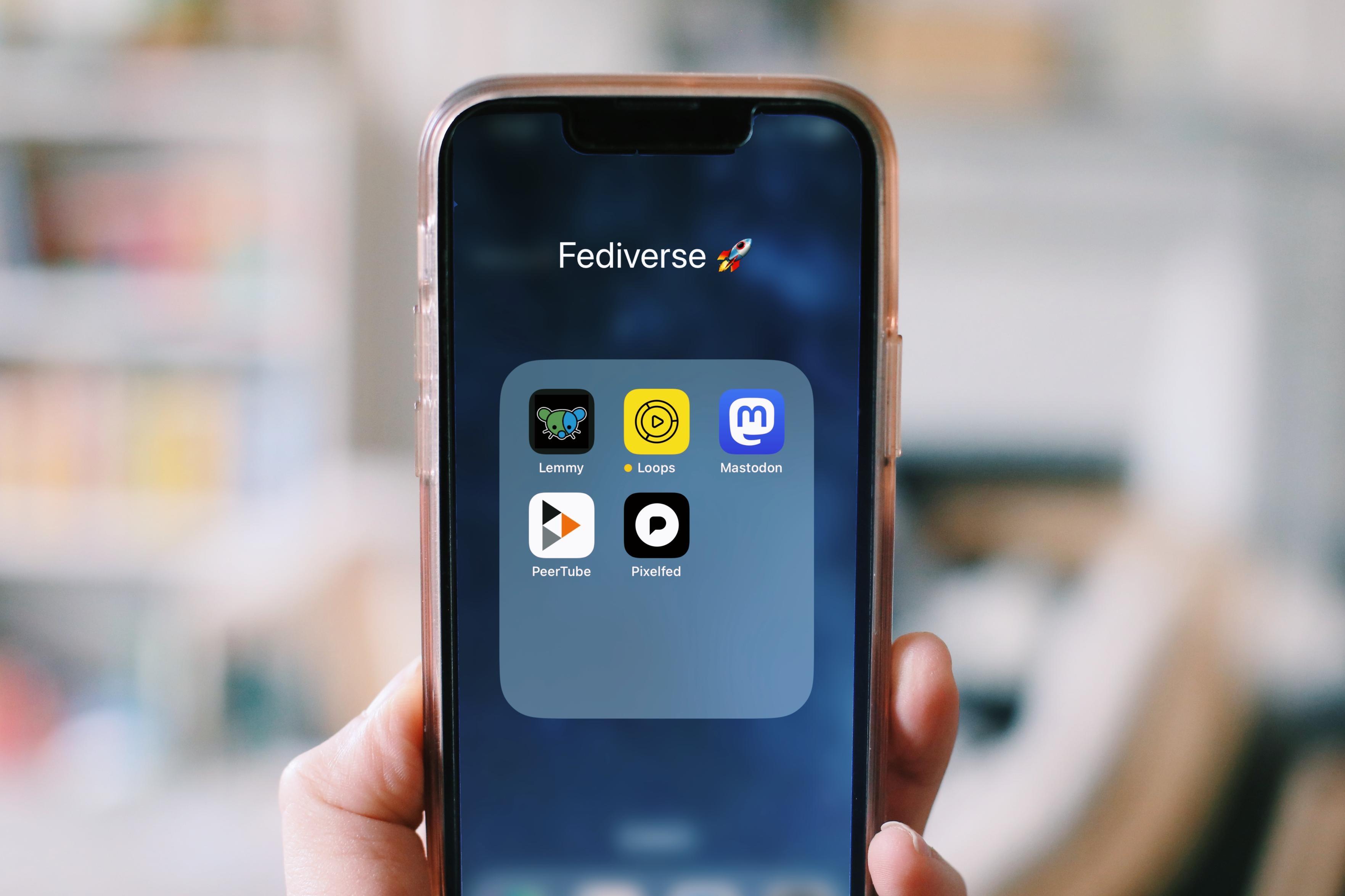 a photo of a hand holding up a smartphone. On screen you see the folder "Fediverse" and 5 app icons: Lemmy (actually a shortcut to the website), Loops, Mastodon, PeerTube and Pixelfed